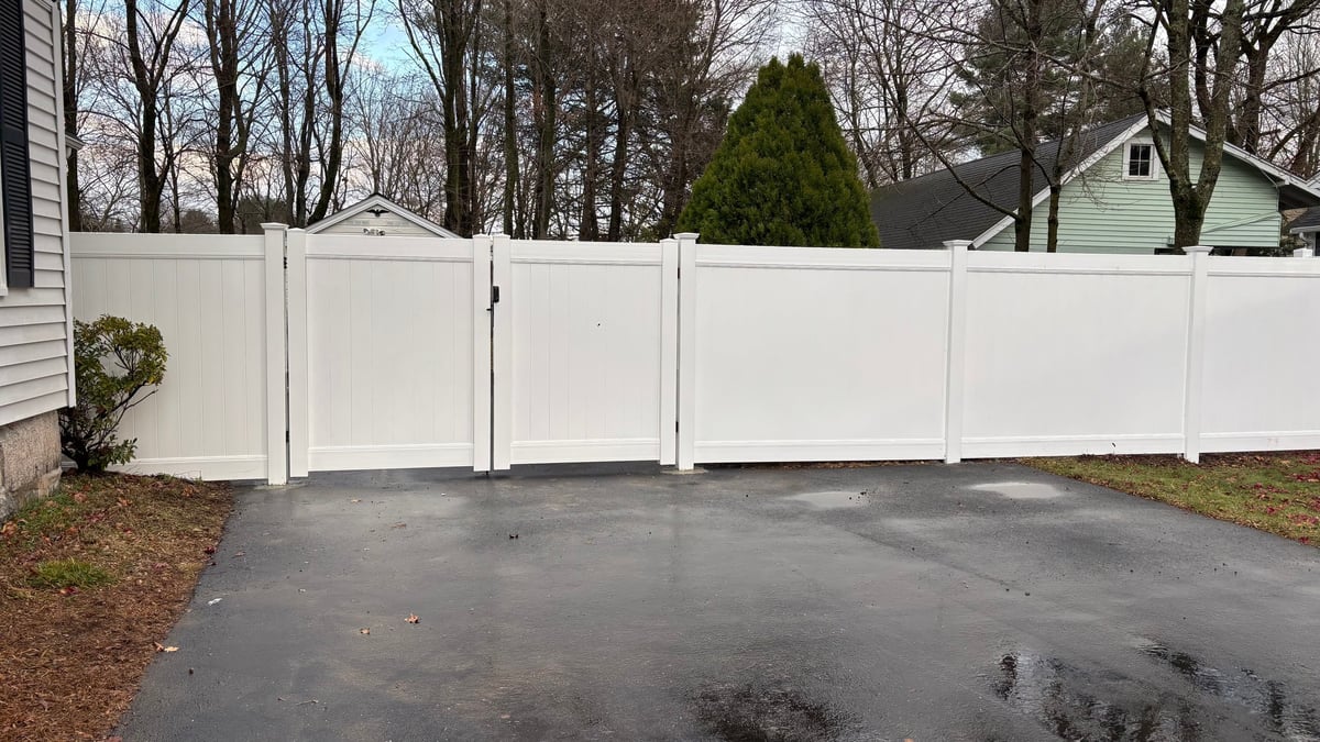 White vinyl privacy fence with gate across a wet driveway in front of residential home with bare trees