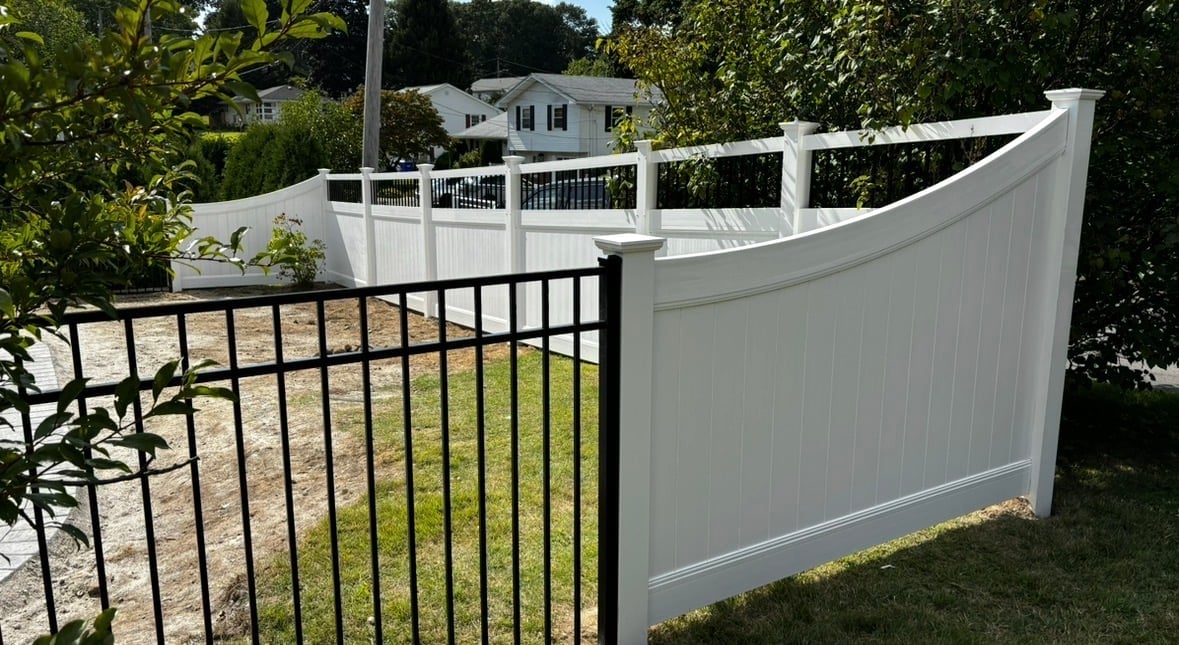White vinyl fence with black metal gate section alongside green lawn and trees, residential home visible in background