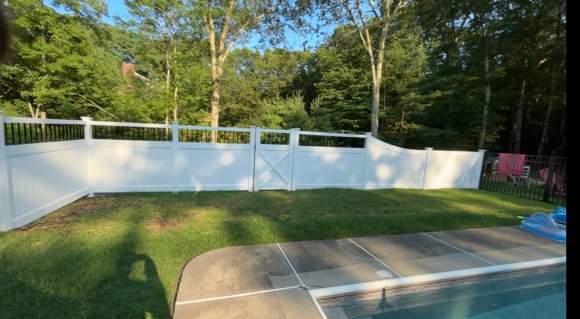 Backyard with white vinyl fence, grassy lawn, concrete patio, and partial swimming pool surrounded by tall trees