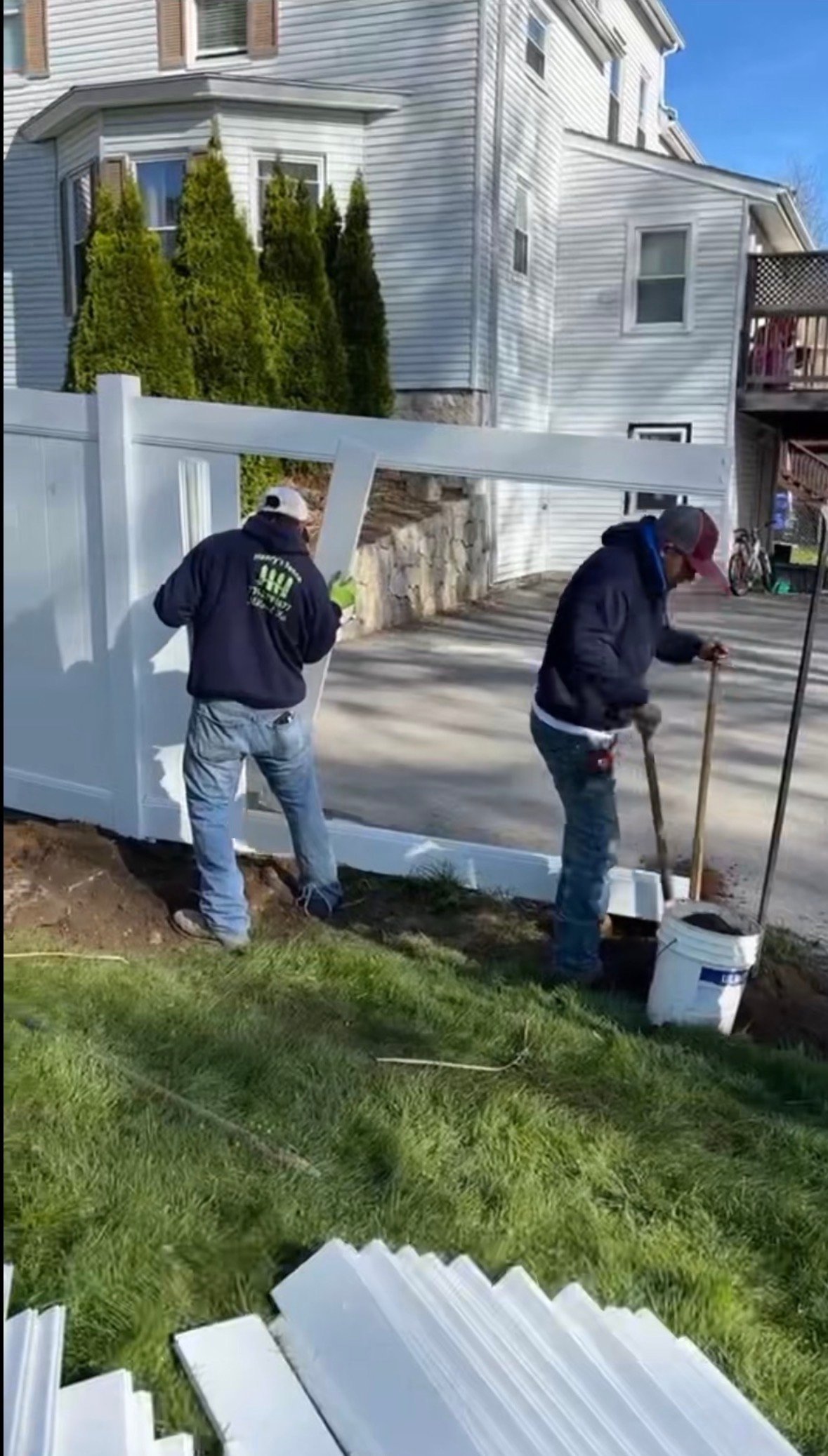 Two workers installing a white vinyl fence in a residential yard with white-sided houses in the background
